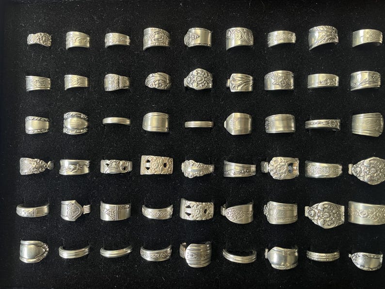 May include: A collection of silver rings displayed on a black surface. The rings vary in design, including plain bands, textured patterns, and floral motifs. The rings are arranged in neat rows, showcasing the variety of styles.