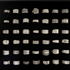 May include: A collection of silver rings with various designs, including floral patterns, geometric shapes, and textured surfaces. The rings are displayed on a black surface, showcasing their intricate details and craftsmanship. The rings vary in size and style.