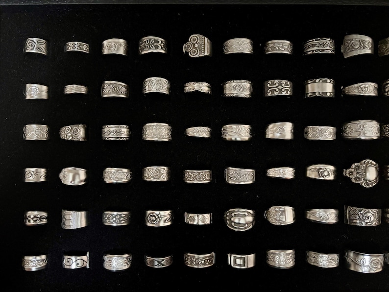 May include: A collection of silver rings with various designs, including floral patterns, geometric shapes, and textured surfaces. The rings are displayed on a black surface, showcasing their intricate details and craftsmanship. The rings vary in size and style.