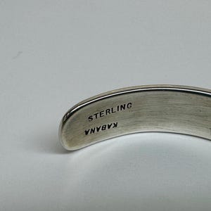 May include: A curved, silver-toned bracelet with the word "STERLING" and the name "KABANA" engraved on the inside. The bracelet has a smooth, polished finish and is set against a light background.