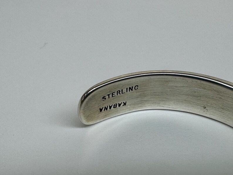 May include: A curved, silver-toned bracelet with the word "STERLING" and the name "KABANA" engraved on the inside. The bracelet has a smooth, polished finish and is set against a light background.