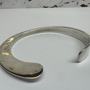 May include: A silver-toned cuff bracelet with a curved, open design. The bracelet has a smooth, polished finish and a rounded, open end. The cuff is a simple, elegant piece of jewelry.