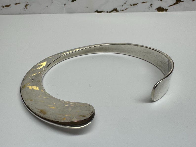 May include: A silver-toned cuff bracelet with a curved, open design. The bracelet has a smooth, polished finish and a rounded, open end. The cuff is a simple, elegant piece of jewelry.