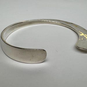 May include: A silver cuff bracelet with a flat, open design. The bracelet has a smooth, polished finish and a curved shape. The opening is approximately 1 inch wide. The bracelet is a simple, elegant piece of jewelry.