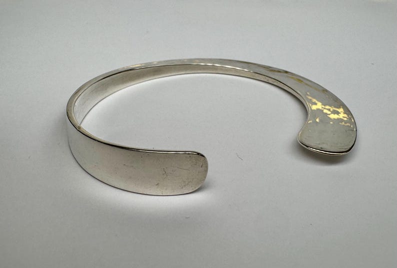 May include: A silver cuff bracelet with a flat, open design. The bracelet has a smooth, polished finish and a curved shape. The opening is approximately 1 inch wide. The bracelet is a simple, elegant piece of jewelry.