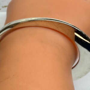 May include: A silver-colored bangle bracelet with a curved design. The bracelet is worn on a wrist, and the metal reflects light. The bracelet appears to be made of a solid, polished metal.