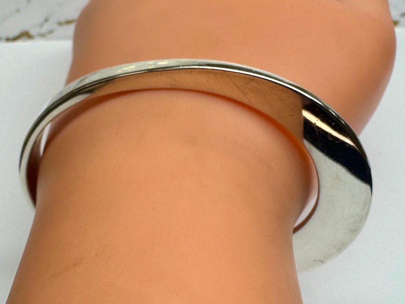 May include: A silver-colored bangle bracelet with a curved design. The bracelet is worn on a wrist, and the metal reflects light. The bracelet appears to be made of a solid, polished metal.