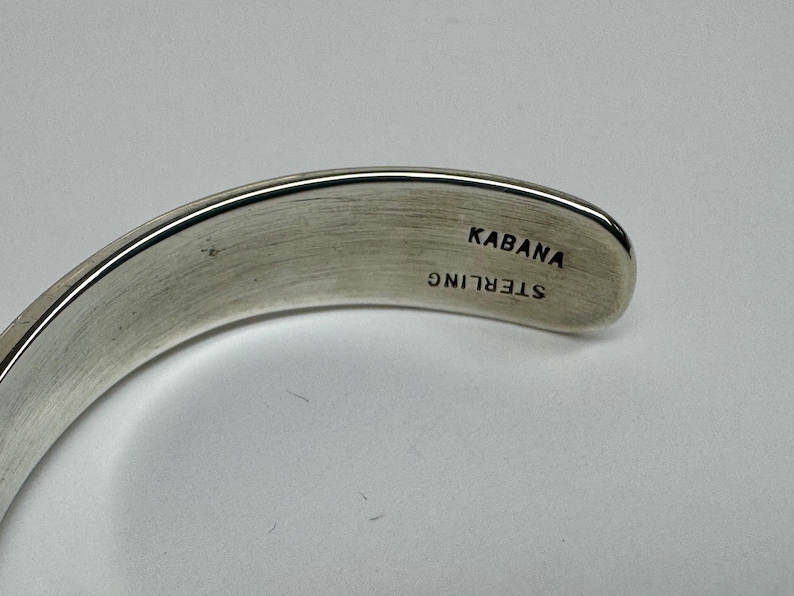 May include: A curved, silver-toned bracelet. The inside of the bracelet is stamped with the word "KABANA" above "STERLING". The bracelet has a smooth, polished finish and is set against a light background.
