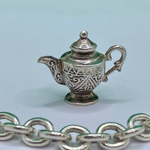 May include: A silver teapot charm with an ornate design. The charm is detailed with intricate floral patterns and a delicate spout.