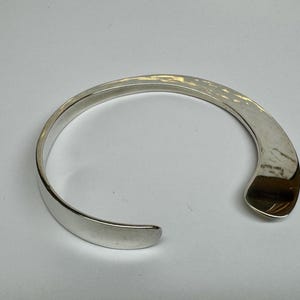 May include: A silver-colored cuff bracelet with a hammered texture on the top surface. The open-ended design allows for easy wear. The bracelet has a smooth, polished finish on the inside and edges. A simple, elegant piece of jewelry.