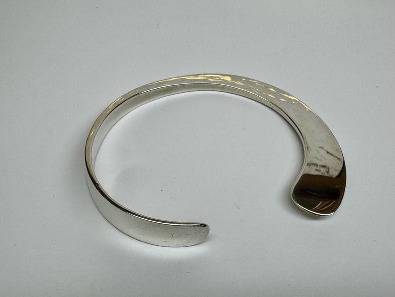 May include: A silver-colored cuff bracelet with a hammered texture on the top surface. The open-ended design allows for easy wear. The bracelet has a smooth, polished finish on the inside and edges. A simple, elegant piece of jewelry.