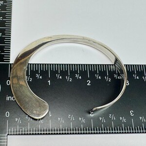 May include: A silver-colored cuff bracelet with a curved design. The open-ended bracelet is placed on a ruler, showing measurements in inches. The bracelet's width is approximately 1 inch.