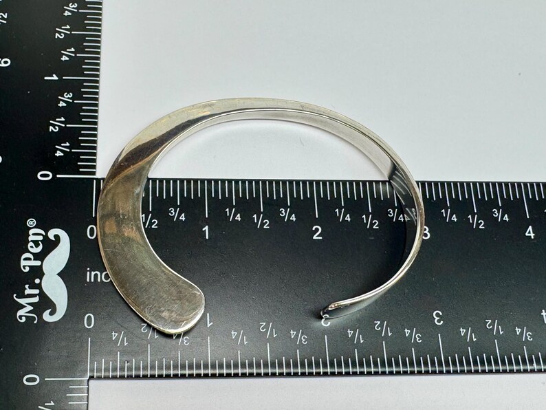 May include: A silver-colored cuff bracelet with a curved design. The open-ended bracelet is placed on a ruler, showing measurements in inches. The bracelet's width is approximately 1 inch.