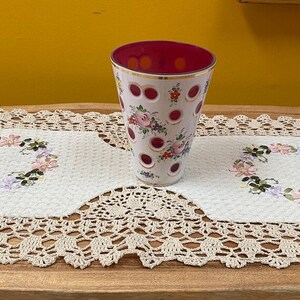 Puede incluir: Un jarrón de vidrio blanco con un borde rojo y un patrón floral sobre un camino de mesa de crochet blanco. El jarrón tiene un patrón de puntos y está decorado con flores rojas, rosas y amarillas.