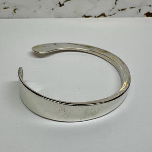 May include: A silver-colored cuff bracelet with a curved, open design. The bracelet has a smooth, polished surface and a simple, elegant style. The open design allows for easy wear and removal. The bracelet is a classic piece of jewelry.