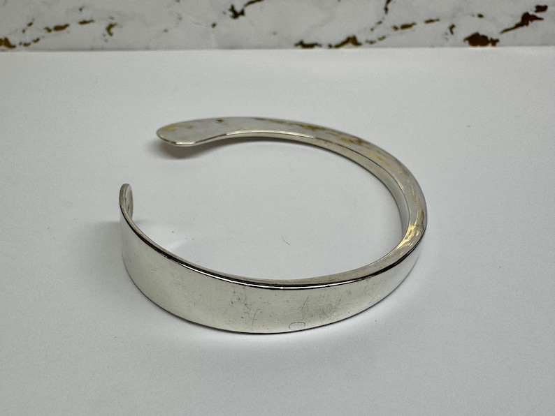 May include: A silver-colored cuff bracelet with a curved, open design. The bracelet has a smooth, polished surface and a simple, elegant style. The open design allows for easy wear and removal. The bracelet is a classic piece of jewelry.