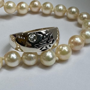 May include: A silver ring with a single clear stone and a design on the band. The ring is resting on a strand of white pearls.