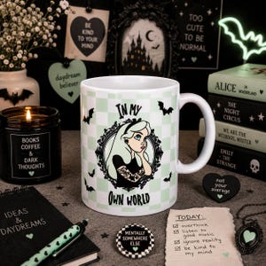May include: A white ceramic mug featuring a light green and white chequered pattern, a central illustration of a person with long hair and tattoos inside an ornate black frame, and the text IN MY OWN WORLD printed in black lettering.