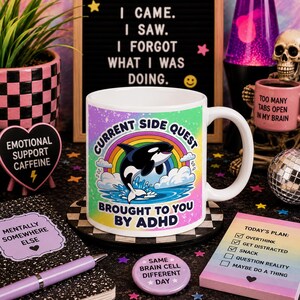 May include: A white ceramic mug featuring a colourful graphic of an orca jumping over a rainbow above blue water. The text reads CURRENT SIDE QUEST BROUGHT TO YOU BY ADHD. The design includes a splatter pattern background in shades of pink, yellow, and blue.