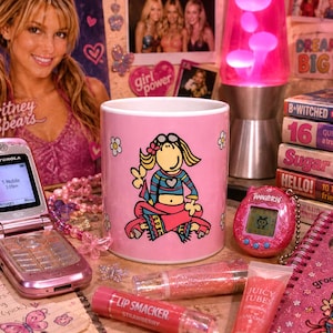 May include: A pink ceramic mug featuring a cartoon character illustration, surrounded by a pink Motorola flip phone, a pink Tamagotchi, lip gloss tubes, a pink notebook, and stacked magazines with text including B*Witched and Sugar.