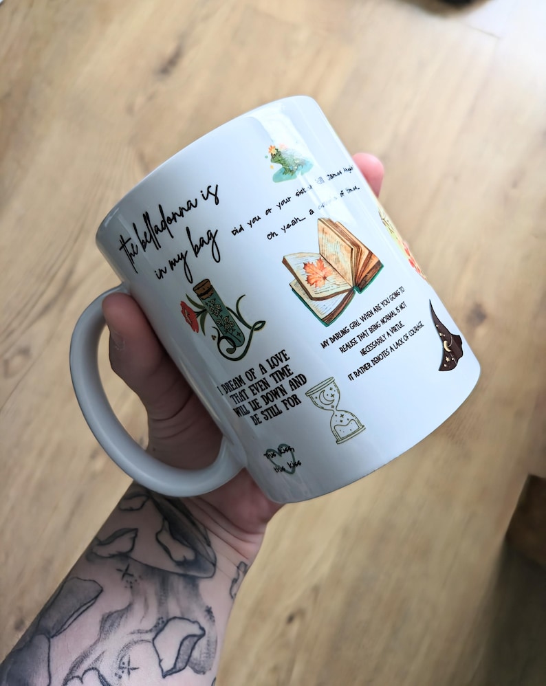 Puede incluir: Una taza de cer&aacute;mica blanca con varias ilustraciones coloridas y texto en negro. El texto dice: The belladonna is in my bag, did you or your sister, oh yeah, a matter of time, I dream of a love that even time will lie down and be still for.