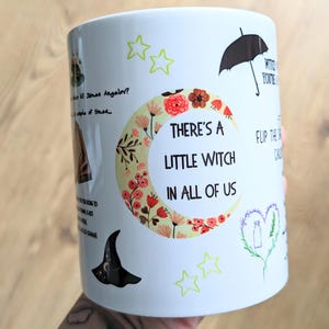 Puede incluir: Una taza de cer&aacute;mica blanca con un gr&aacute;fico de luna creciente lleno de patrones florales y el texto "THERE'S A LITTLE WITCH IN ALL OF US". Las ilustraciones adicionales incluyen un sombrero de bruja negro, un paraguas, estrellas y citas.