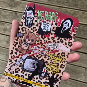 May include: A rectangular sticker sheet with a leopard print background featuring horror-themed illustrations including a mobile phone, a ghost mask, a teddy bear with an axe, a television, and text reading Horror, You've Got A Curse, Certified Screamer, Final Girl Entry, Slay The Day, Just One More Scream, Emotionally Attached To Fictional Killers, and Crime Scene - Do Not Cross.