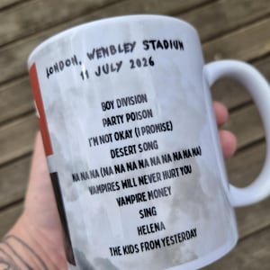 May include: A white ceramic mug featuring a grey marbled background and black text listing concert details: London, Wembley Stadium, 11 July 2026, followed by a tracklist including Boy Division, Party Poison, I'm Not Okay (I Promise), and others.