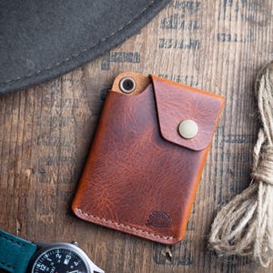 May include: A brown leather wallet with a snap closure and a metal eyelet. The wallet is on a wooden surface with a watch, a hat, and a rope. The watch has a green strap and a silver face.