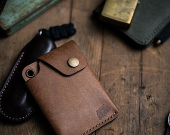 May include: A tan leather wallet featuring a single brass snap button closure, a reinforced metal eyelet on the top left corner, and a stamped logo in the bottom right corner. The wallet is constructed with visible stitching along the edges.