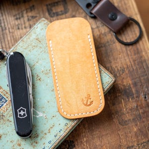 Könnte beinhalten: Eine Auswahl an Lederaccessoires, darunter eine hellbraune Lederscheide mit Anker-Design, ein schwarzes Multitool-Messer und ein brauner Leder-Schlüsselanhänger. Die Artikel liegen auf einer rustikalen Holzoberfläche.