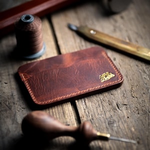 May include: A brown leather wallet with gold lettering reading "The Leather", alongside leatherworking tools such as a hammer, awl, and thread spool. The wallet is rectangular with rounded corners and stitched edges.