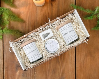 May include: A gift box with candles and a small soap bar. The box contains two glass jar candles with wooden lids, a small tin candle, and a soap bar with a tree design. The candles are labeled "Apple Crumble" and "Autumn Bonfire".