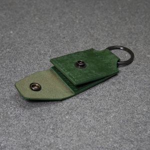 May include: A green leather keychain holder with a snap closure and a metal key ring. The holder is open, revealing a lighter green interior. The leather has a textured appearance.