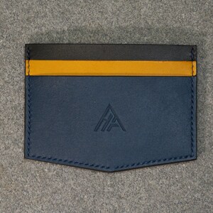 May include: A navy blue leather cardholder with a black and yellow stripe across the top. The cardholder has a stitched border and a debossed logo in the lower center.