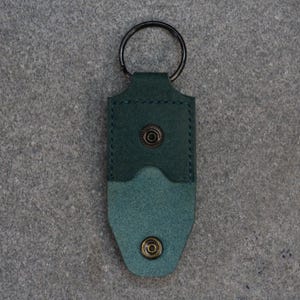 以下が含まれることがあります： 黒い金属リングが付いた、ティール色の革製キーホルダー。ホルダーはツートンカラーのデザインで、濃いティール色のトップ部分と明るいティール色のボトム部分があります。真鍮色のボタンが付いたスナップ留めです。