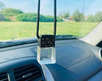 May include: A black and gold car air freshener hanging from the rearview mirror of a car. The air freshener is a small square bottle with a decorative metal cage around it.