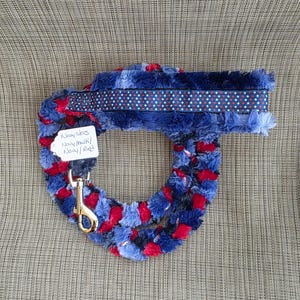 May include: A pet leash made of plush, textured fabric in navy blue, light blue, and red, featuring a silver metal swivel snap hook. A navy blue ribbon with a white star and red dot pattern runs along the top. A white tag reads Navy stars Navy/multi/Navy/Red.