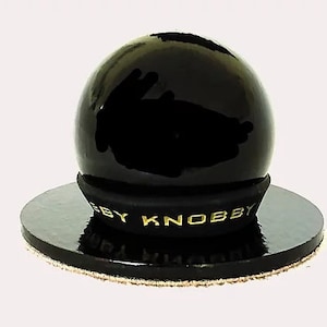 May include: A black, glossy, spherical knob with a gold "BY KNOBBY" inscription on the base. The knob is sitting on a black circular base with a brown fabric edge.