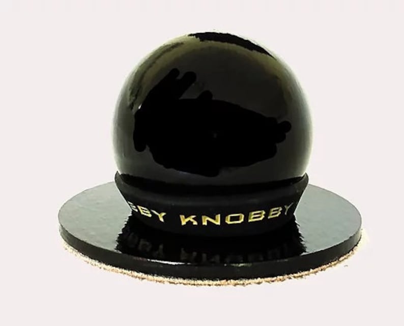 May include: A black, glossy, spherical knob with a gold "BY KNOBBY" inscription on the base. The knob is sitting on a black circular base with a brown fabric edge.