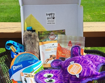 May include: A yellow box filled with dog toys and treats. The box has a label that says "happy mail" and "natural dawg". The toys include a purple squeaky toy, a blue and orange frisbee, a brown and white plush toy, and a blue and white rope toy. The treats are in a bag that says "small good for small & smaller made with chicken".