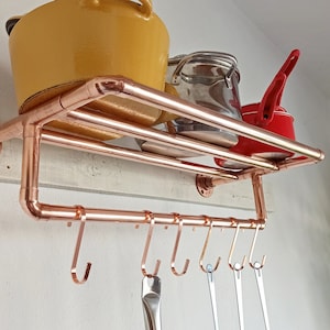 Op de afbeelding: Koperen pijp-rek met haken om keukengerei aan te hangen. Het rek heeft twee niveaus en is gemonteerd aan een witte muur. Er staan drie potten op het rek, een gele, een zilveren en een rode.