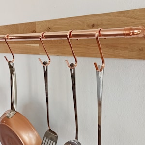 Könnte beinhalten: Ein Kupferrohrregal mit Haken, das an einer Holzplatte befestigt ist. Das Regal hält verschiedene Küchenutensilien, darunter einen Kupfertopf, einen Spatel, eine Schöpfkelle und ein Sieb.