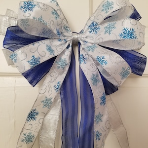 May include: A large blue and white bow with a snowflake pattern. The bow is made of ribbon and mesh and has a silver glitter finish.