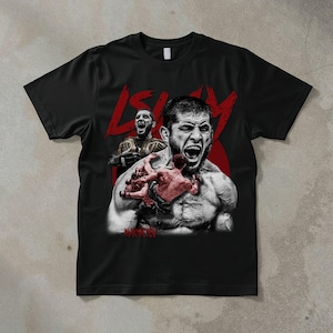 May include: Black t-shirt featuring a graphic of a mixed martial artist in a fighting pose. The design includes a red stylized text and images of the fighter with championship belts. The shirt is made of a soft material.