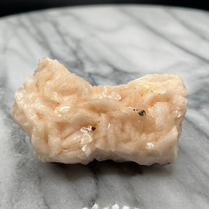 Puede incluir: Un espécimen mineral rosa y blanco con una superficie rugosa y texturizada. El mineral está sobre una superficie de mármol blanco.