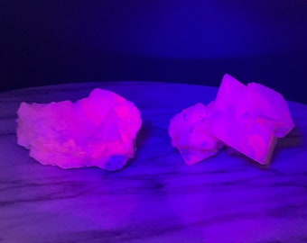 Puede incluir: Dos grupos de cristales rosas brillando bajo una luz negra. Los cristales están sobre una superficie de mármol blanco.