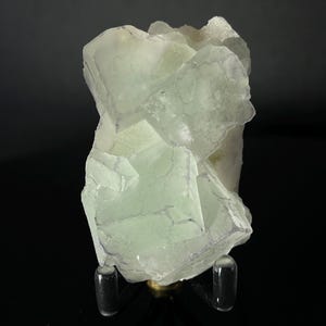 Peut inclure: Un grand amas de cristaux de fluorine vert clair avec une légère teinte violette. Le cristal est présenté sur un support noir avec trois pieds transparents.