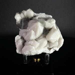 Peut inclure: Un amas de cristaux blancs et gris, possiblement du quartz ou un minéral similaire, est présenté sur une surface noire. Les cristaux ont une texture rugueuse et irrégulière. Le spécimen minéral est monté sur un support transparent.