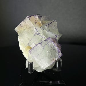 Peut inclure: Un groupe de cristaux de fluorine vert pâle avec des bandes violettes. Les cristaux sont disposés en forme de pyramide et sont exposés sur un support noir.